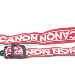 Op de afbeelding: Twee rode Canon camerariemen met witte tekst en zwarte plastic clips. De riemen liggen op een wit oppervlak. Het woord "CANON" wordt herhaald over de lengte van de riemen.