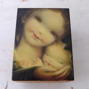 Puede incluir: Una caja de música de madera vintage con una imagen pintada de una mujer sosteniendo un bebé dormido. La mujer tiene el pelo largo y castaño y lleva un vestido oscuro. El bebé está envuelto en una manta blanca.