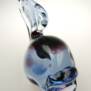 May include: A blue and purple glass sculpture of a fish with a curled tail. The fish has a white stripe on its side and a pink mouth.