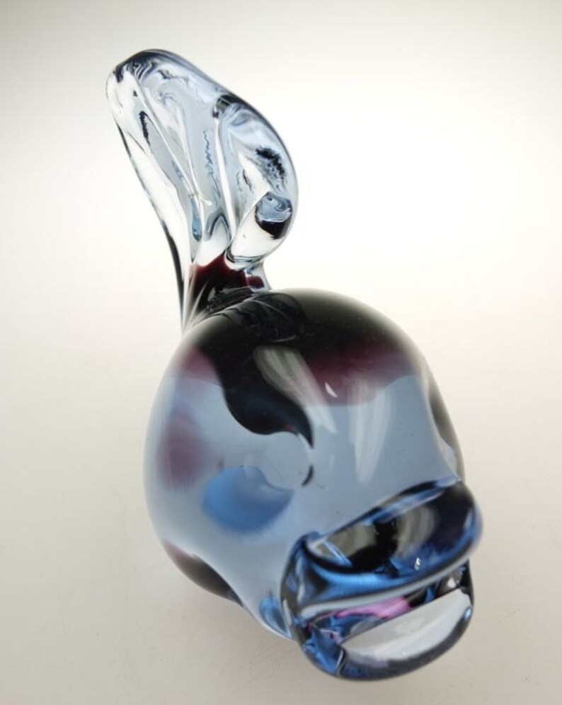 May include: A blue and purple glass sculpture of a fish with a curled tail. The fish has a white stripe on its side and a pink mouth.