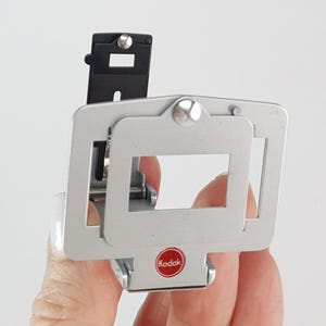 Könnte beinhalten: Silberner Diaprojektor mit einem schwarzen Diahalter und einem roten Kodak-Logo.