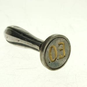 May include: A silver seal with a handle. The seal has a circular face with the letters "E.O." in gold lettering.