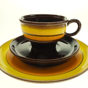 Könnte beinhalten: Ein braunes und gelbes Keramik-Teetassen- und Untertassen-Set. Die Tasse hat einen breiten Rand und einen Henkel. Die Untertasse hat einen gelben Rand und eine braune Mitte.