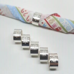 May include: Six silver napkin rings with an ornate design. The rings are arranged in a row on a white surface.