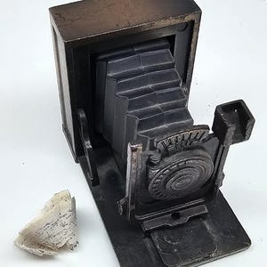 Może przedstawiać: Ciemnobrązowa figurka aparatu mieszkowego w stylu vintage, wykonana z litego materiału. Aparat posiada prostokątny korpus, harmonijkowy mieszek oraz okrągły obiektyw. Obok znajduje się mały, fakturowany kawałek kory brzozowej.