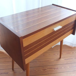 May include: A mid-century wooden cabinet with a drawer and tapered legs. The cabinet has alternating bands of light and dark wood, with a light-coloured handle on the drawer. The top has inlaid light wood stripes.