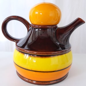 Könnte beinhalten: Eine braune und orangefarbene Teekanne aus Keramik mit einem runden, orangen Deckel. Die Teekanne hat einen gelben Streifen um die Mitte und einen braunen Henkel.
