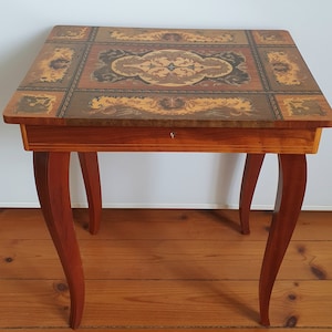 Könnte beinhalten: Kleiner, quadratischer Holztisch mit eingelegter Tischplatte. Die Platte zeigt ein zentrales Blumendesign in Braun-, Beige- und Schwarztönen. Er hat eine Schublade und geschwungene Beine, im klassischen, antiken Stil.