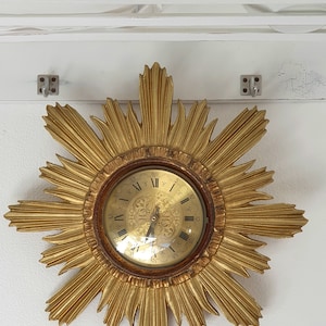 Puede incluir: Un reloj de sol dorado con una esfera redonda y adornada. La esfera del reloj tiene números romanos y un diseño floral detallado.