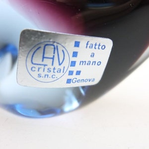 May include: A clear glass paperweight with a blue and purple ombre effect. The paperweight has a sticker on the side that reads "CAV Cristal S.N.C. fatto a mano Genova".