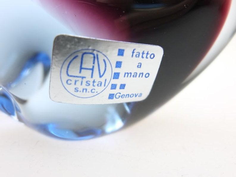 May include: A clear glass paperweight with a blue and purple ombre effect. The paperweight has a sticker on the side that reads "CAV Cristal S.N.C. fatto a mano Genova".