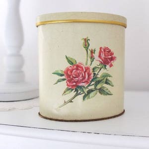 May include: A vintage cream-coloured metal canister with a gold trim and a floral design featuring pink roses.