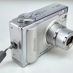 May include: A silver Fujifilm FinePix F10 digital camera featuring a Fujinon zoom lens, a top-mounted power button, and a shutter release. The camera body includes the FinePix logo, a 6.3 megapixels label, and an attached grey wrist strap.
