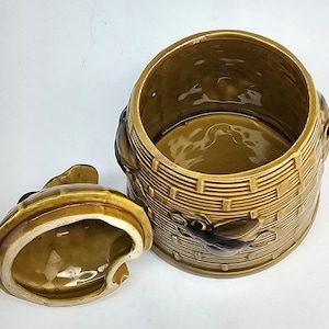 May include: A cylindrical ceramic honey pot with a glossy mustard yellow glaze and a textured basket-weave pattern. The jar features a raised, dark brown bee motif on the side and includes a matching removable lid with a small bee-shaped handle.