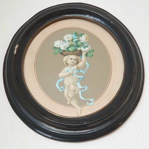 Könnte beinhalten: Gerahmtes Kunstwerk mit einer cherubischen Figur, die einen Korb mit weißen Rosen trägt. Der Cherub ist mit blauen Bändern geschmückt, vor einem neutralen Hintergrund und in einem schwarzen, runden Rahmen. Das Kunstwerk hat eine klassische, romantische Ästhetik.
