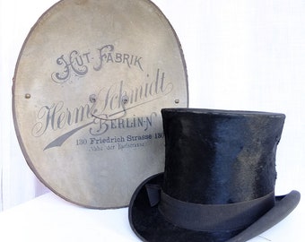 May include: A black top hat with a black band sits on a white surface next to a round, cream-colored sign with black text that reads "Hut Fabrik Herm. Schmidt Berlin-N 130 Friedrich Strasse 130 Nahe der Karlstrasse."