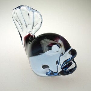 May include: A glass figurine of a fish with a blue, white, and black swirl pattern. The fish has a large, open mouth and a tail that is curled up.
