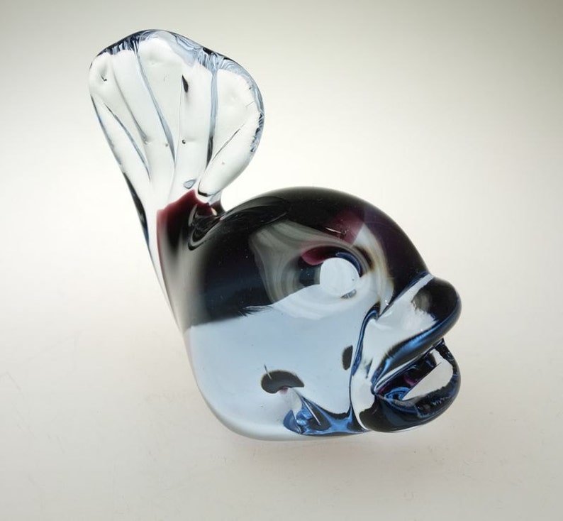 May include: A glass figurine of a fish with a blue, white, and black swirl pattern. The fish has a large, open mouth and a tail that is curled up.