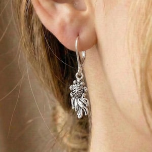 May include: Silver-coloured dangle earring with a detailed, stylised fish design. The earring hangs from a simple hook, displaying the intricate details of the fish's scales and fins. The earring is worn on a person's ear.