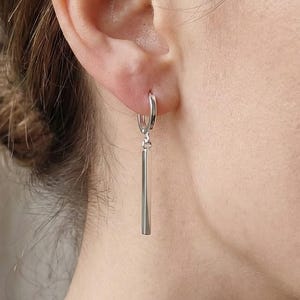 Puede incluir: Primer plano de un pendiente de aro plateado con un colgante largo y rectangular. El pendiente se lleva en la oreja. El fondo es de color neutro. Diseño minimalista.