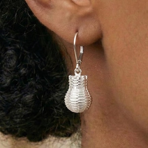 May include: A silver earring with a basket design is displayed. The earring hangs from a lever-back clasp. The basket-shaped charm has a textured, woven appearance. The earring is shown against a blurred background. The earring is made of silver.