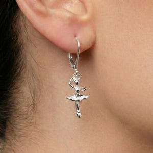 Könnte beinhalten: Silberne Ohrhänger mit Ballerina-Motiv. Die Ohrringe zeigen eine kleine Ballerina-Figur mit Tutu und erhobenen Armen. Die Ohrringe sind an einem silbernen Haken befestigt.
