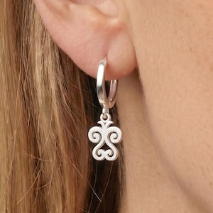 Peut inclure: Boucle d'oreille argentée avec un pendentif décoratif. Le pendentif présente un motif symétrique avec des lignes courbes et des volutes. La boucle d'oreille est portée à l'oreille d'une personne, l'accent étant mis sur le bijou.
