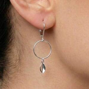 Puede incluir: Pendientes colgantes plateados con un diseño de aro circular y un pequeño dije en forma de hoja. Los pendientes están hechos de un material metálico brillante y se muestran puestos en una oreja. Diseño simple y elegante.