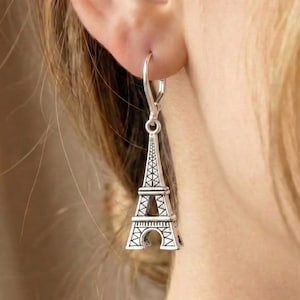 May include: Silver Eiffel Tower drop earring. The earring hangs from a lever-back clasp. The Parisian landmark is depicted with detailed openwork and a textured finish. The earring is worn on the ear.