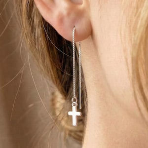 May include: A silver threader earring featuring a small, polished cross charm suspended from a delicate chain. The earring is designed to be pulled through the earlobe, allowing the chain to hang down with the cross charm positioned at the end.