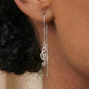 Puede incluir: Un pendiente de hilo de plata con un dije de clave de sol. El pendiente consiste en una delicada cadena de eslabones cuadrados que atraviesa el lóbulo de la oreja, con el símbolo musical colgando de un extremo de la cadena.