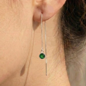 Pode incluir: Um brinco de prata com uma pequena pedra preciosa redonda verde esmeralda. O brinco pende de uma delicada corrente que passa pela orelha. O brinco é mostrado em close-up.