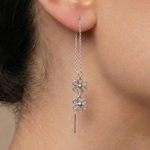 Può includere: Orecchini pendenti color argento con due delicati ciondoli a forma di fiore e una barra verticale. Gli orecchini sono sospesi a una catena sottile, creando un design aggraziato e fluente. Gli elementi floreali aggiungono un tocco di eleganza ispirata alla natura.