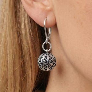 May include: A silver dangle earring featuring a spherical charm with an intricate, openwork filigree pattern. The charm hangs from a simple silver hoop wire that passes through the earlobe, showcasing a polished metallic finish and dark recessed details.