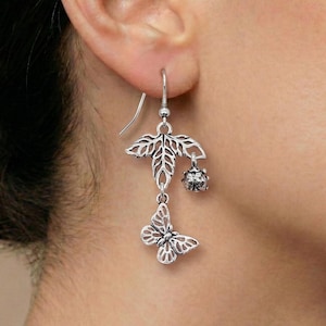 Puede incluir: Pendientes colgantes plateados con una hoja, un pequeño dije de cuentas y una mariposa. Los pendientes están hechos de un metal brillante y se usan en la oreja. El diseño es intrincado y detallado, mostrando un tema inspirado en la naturaleza.