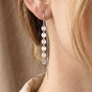 May include: A silver chain earring with six small, round, flat charms hanging from the ear. The earring is worn by a person with blonde hair and a beige top. The earring is a simple, elegant design.