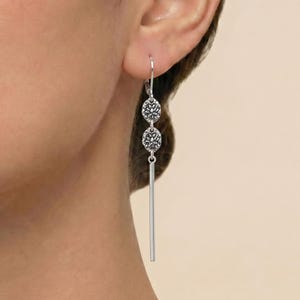 May include: A silver drop earring featuring two small, circular, textured floral-patterned charms connected vertically, ending in a thin, smooth, straight silver bar that hangs downward from the earlobe on a simple silver hook wire.
