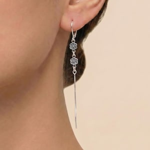 Könnte beinhalten: Silberne Hängeohrringe mit drei kleinen Blumenanhängern und einem langen, schlanken Stab. Die Ohrringe sind so konzipiert, dass sie vom Ohr hängen und einen zarten und eleganten Look erzeugen. Die Blumen haben eine dunkelgraue Mitte.