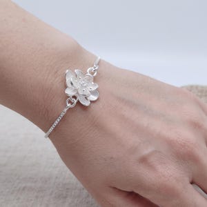 May include: A silver-toned bracelet featuring a central floral charm shaped like a lotus blossom. The bracelet consists of a delicate chain that connects to the flower, which has layered petals and a textured center, creating a metallic finish.