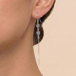 May include: Silver drop earrings featuring three small, gray flower charms. The earrings have a long, slender, silver bar that hangs down. The earrings are shown on a person's ear.
