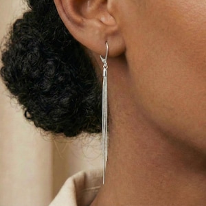 Puede incluir: Pendientes colgantes plateados con un diseño de cadena en cascada. Los pendientes están sujetos a un cierre de palanca y cuelgan hacia abajo, creando un efecto fluido. Los pendientes se muestran en una oreja.