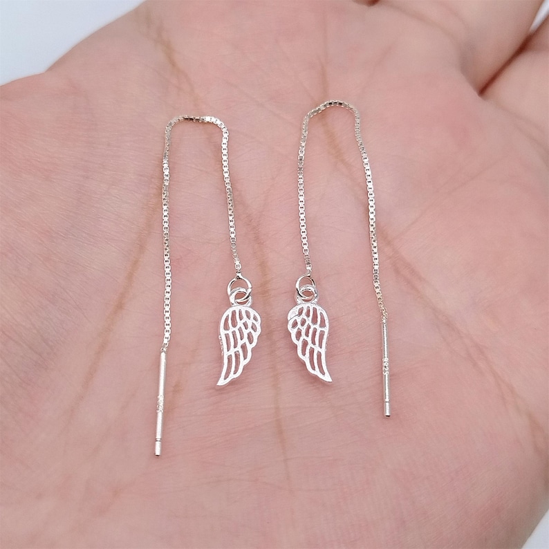 May include: A pair of silver threader earrings. Each earring has an openwork wing charm hanging from a fine chain. The chain extends upwards and ends with a straight bar. The earrings are shown against a neutral background.