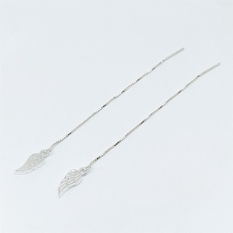 May include: A pair of silver threader earrings. Each earring has a small, detailed wing charm at the base of a delicate chain. The earrings are shown against a plain white background, highlighting their elegant design.