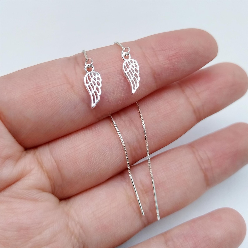 May include: A pair of silver threader earrings. Each earring has a small, open-winged charm hanging from a fine chain. The wing charms are detailed with lines. The earrings are shown against a light background.