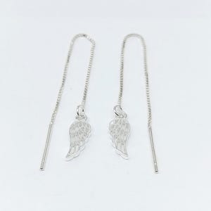 May include: A pair of silver threader earrings. Each earring has a small wing charm hanging from a fine chain. The earrings are designed to be pulled through the ear, creating a simple and elegant look. The background is plain white.