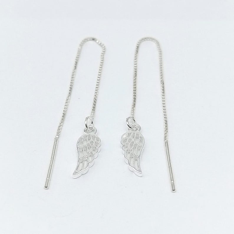 May include: A pair of silver threader earrings. Each earring has a small wing charm hanging from a fine chain. The earrings are designed to be pulled through the ear, creating a simple and elegant look. The background is plain white.