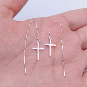 Puede incluir: Un par de pendientes de cruz plateados. Cada pendiente presenta un pequeño colgante de cruz suspendido de una delicada cadena. Los pendientes están diseñados para pasar por la oreja, creando un look sencillo pero elegante.
