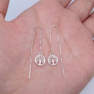 May include: A pair of silver threader earrings. Each earring features a small, round charm with a cut-out tree design. The earrings are displayed against a neutral background.