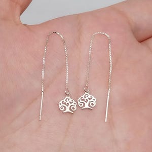 Könnte beinhalten: Ein Paar silberne Ohrringe mit einem Baumdesign. Die Ohrringe bestehen aus einer zarten Kette und haben einen kleinen Baum-Anhänger am unteren Ende.