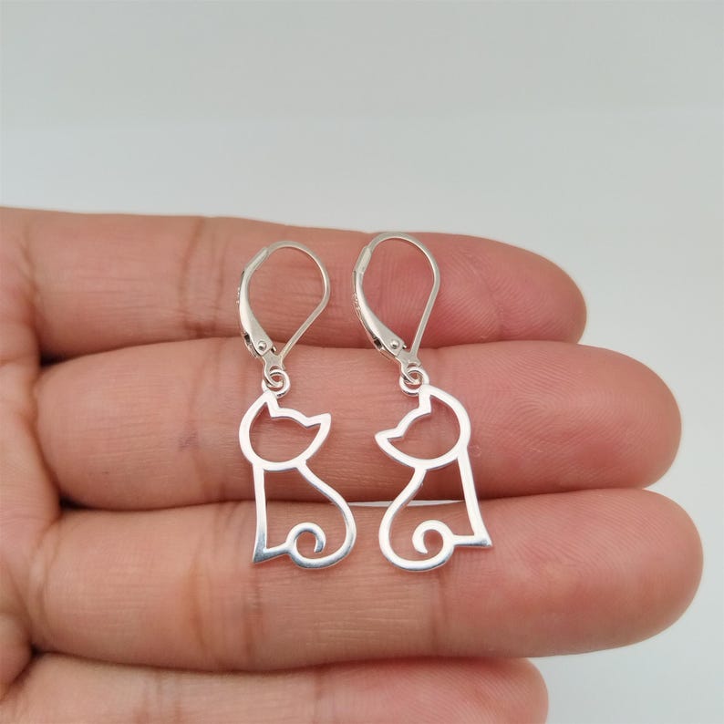 May include: A pair of silver dangle earrings featuring a cutout design of two cats facing each other.